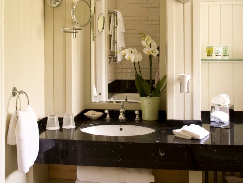 Een badkamerblad heeft een zwart marmeren oppervlak met een witte wasbak. Twee heldere glazen en handdoeken staan bij de wasbak. Een groene orchidee in een pot met witte bloemen staat bij de spiegel. Toiletartikelen aan de muur en een ronde vergrootspiegel zijn zichtbaar. Subtiele verlichting versterkt de scène. | Echt Ierland Vakanties