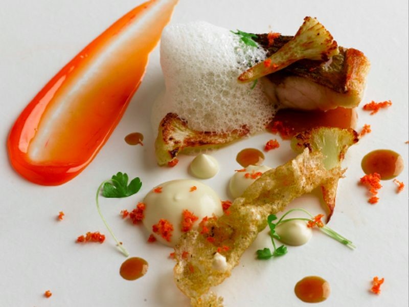 Gourmet gerecht met kunstig gerangschikt eten op een wit bord. Inclusief een veeg feloranje saus, wit schuim, knapperig gebruinde vis, kleine druppeltjes beige en bruine saus, groene kruiden en een delicate, gefrituurde garnering, wat een visueel aantrekkelijke compositie oplevert. | Echt Ierland Vakanties