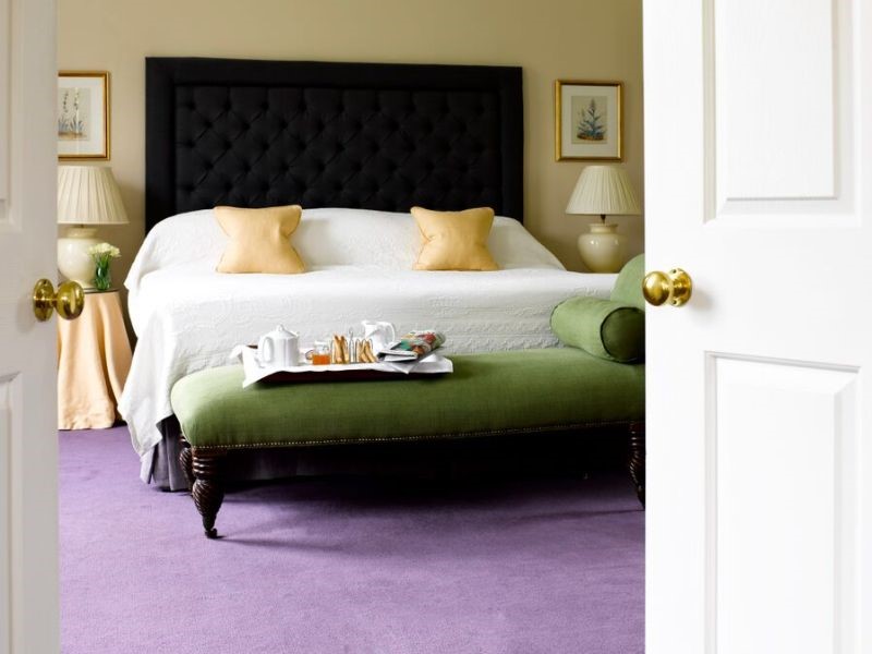 Kijk door open deuren naar een slaapkamer met een groot bed met een zwart, gecapitonneerd hoofdeinde en wit beddengoed met twee gele kussens. Een groene bank aan het voeteneind bevat een ontbijtblad. De kamer heeft een lavendelkleurig tapijt, beige muren en ingelijste kunst boven nachtkastjes met lampen. | Echt Ierland Vakanties
