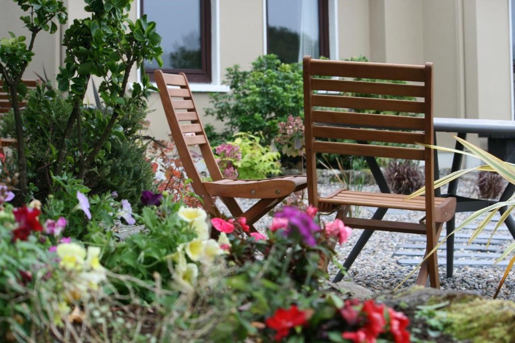 Tuintafereel met twee houten stoelen tegenover elkaar tussen kleurrijke bloemen en weelderig groen. Een met kiezelstenen bedekt pad loopt ertussen. Op de achtergrond is een beige gebouw met ramen gedeeltelijk zichtbaar, wat bijdraagt aan de serene buitenomgeving. | Echt Ierland Vakanties