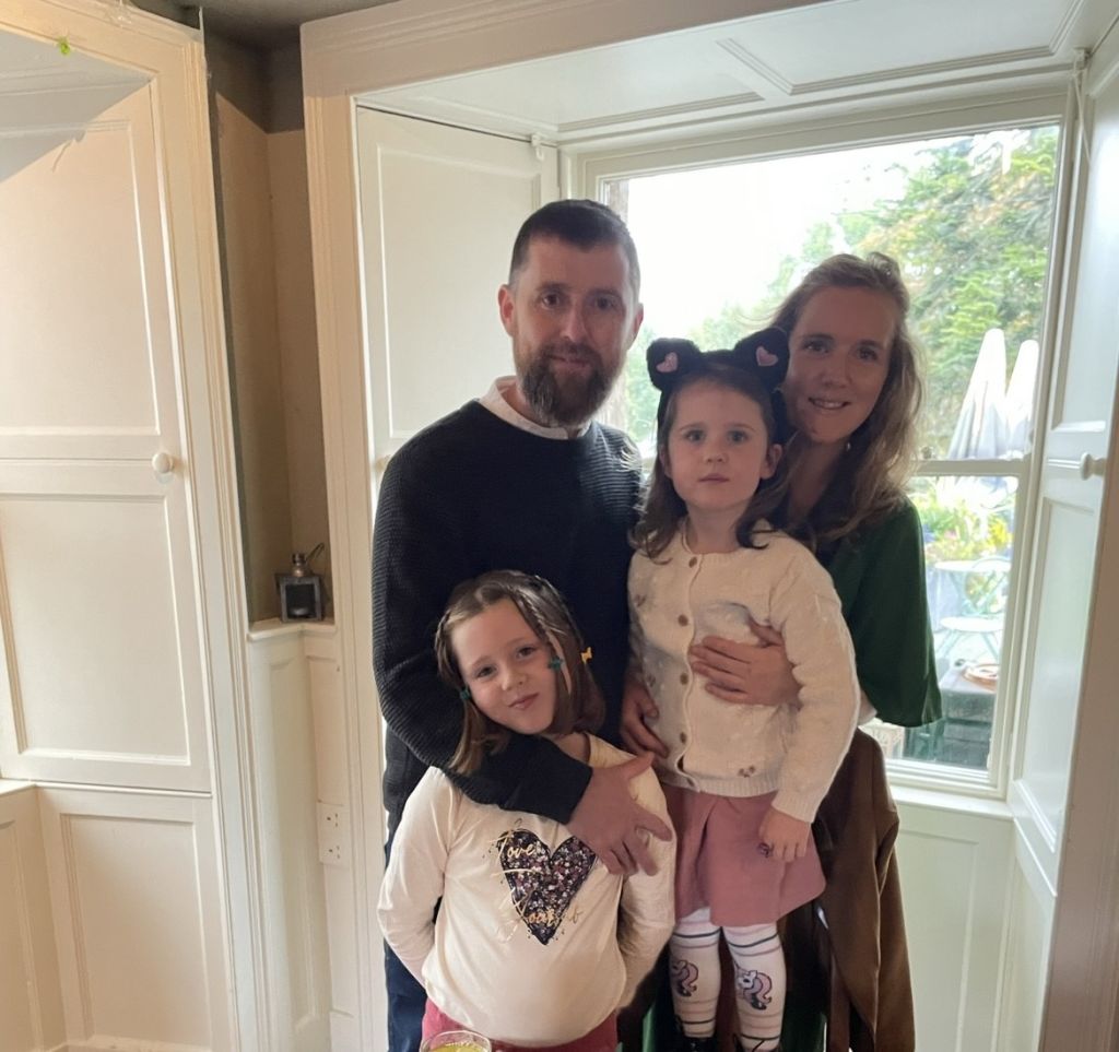 Een gezin van vier verzamelt zich in Hazelwood Lodge bij een groot raam. De man, met een baard in een donkere trui, en de vrouw, met golvend haar in een groen topje, houden hun dochters vast. De een draagt een kattenoor-hoofdband; de ander draagt een topje met hartjesmotief. Buiten maken bomen het serene uitzicht compleet. | Echt Ierland Vakanties