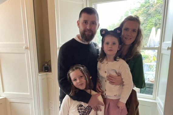 Een gezin van vier verzamelt zich in Hazelwood Lodge bij een groot raam. De man, met een baard in een donkere trui, en de vrouw, met golvend haar in een groen topje, houden hun dochters vast. De een draagt een kattenoor-hoofdband; de ander draagt een topje met hartjesmotief. Buiten maken bomen het serene uitzicht compleet. | Echt Ierland Vakanties