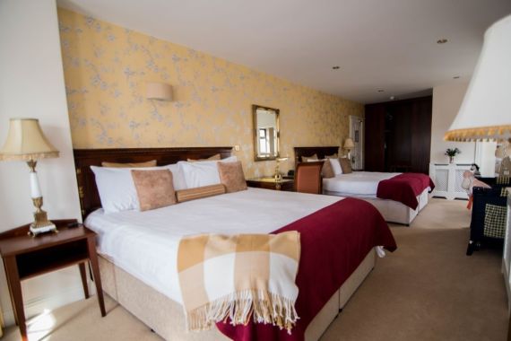 Een gezellige hotelkamer met twee grote bedden, elk versierd met wit linnen, bruine kussens en rode spreien. Een beige en rode plaid deken is gedrapeerd over een bed. Elegante bedlampjes, een bloemenbehang, een spiegel met gouden lijst en een klein bureau maken de warme, uitnodigende ambiance compleet. | Echt Ierland Vakanties