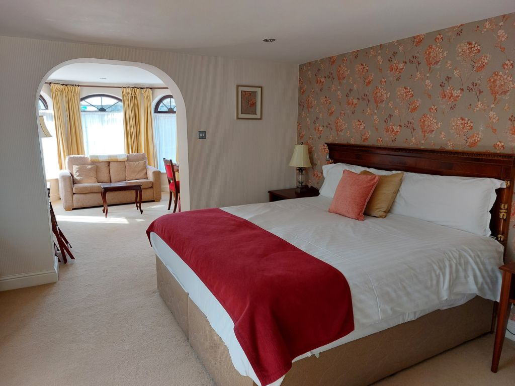 Een gezellige hotelkamer heeft een groot bed met wit linnen en een rode deken. Een oranje bloemenaccentmuur en een klein ingelijst schilderij zijn zichtbaar. Door een boog verschijnt een zithoek met een beige bank, rode stoelen en ramen met gele gordijnen. | Echt Ierland Vakanties