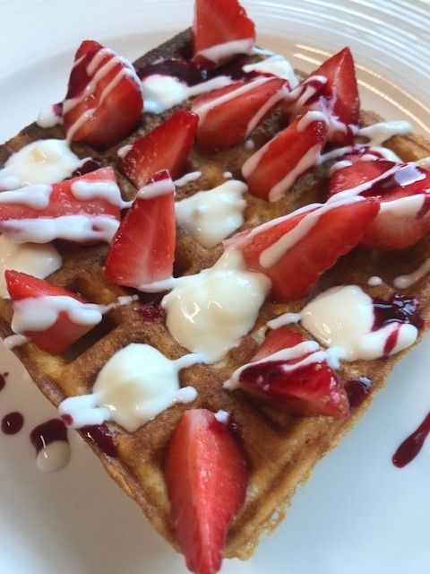 Een gouden wafel met plakjes aardbeien, besprenkeld met witte glazuur en een vleugje bessensaus, geserveerd op een wit bord. De aardbeien zijn netjes gerangschikt en de toppings geven het gerecht een levendige, zoete uitstraling. | Echt Ierland Vakanties