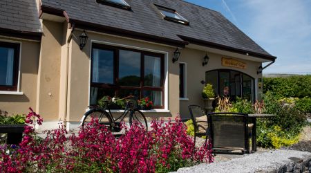 Een charmant huis met een donkergrijs dak, met meerdere dakramen. Een goed onderhouden tuin met levendige roze bloemen en een vintage fiets sieren de voorkant. Een stenen muur grenst aan de tuin en er is een terrasset met twee stoelen en een tafel. Een helderblauwe lucht hangt erboven. | Echt Ierland Vakanties