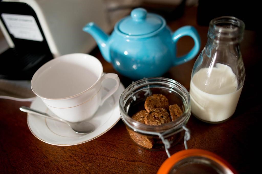 Een blauwe theepot, een witte theekop met een schotel en lepel, een glazen pot met koekjes en een glazen fles melk staan op een houten tafel. De scène suggereert de voorbereiding op theetijd, met de items netjes op hun plaats. | Echt Ierland Vakanties