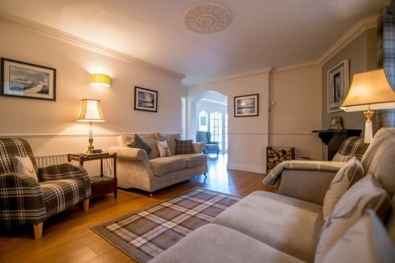 Gezellige woonkamer met twee geruite fauteuils, een beige bank en een geruit kleed op een houten vloer. Zachte verlichting van twee staande lampen en een wandlamp. Ingelijste kunstwerken sieren crèmekleurige muren. Rechts is een open haard zichtbaar en dubbele deuren leiden naar een andere kamer op de achtergrond. | Echt Ierland Vakanties