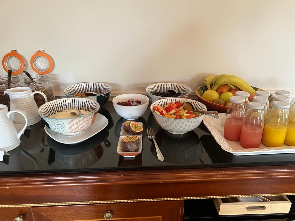 Een ontbijtbuffet met kommen ontbijtgranen, granola, melk en verse fruitsalade. Ook zijn er plakjes passievrucht en potten met zaden aanwezig. Rechts staan flessen met sinaasappel- en roze sappen. Een kom bananen en appels maakt het geheel compleet op een houten dressoir. | Echt Ierland Vakanties