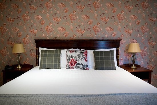 Welkom bij Hazelwood Lodge, waar een gezellige slaapkamer wacht. Het beschikt over een groot bed met een donkerhouten hoofdeinde, versierd met twee plaids en één bloemenkussen. Elk houten nachtkastje heeft een lamp met een beige kap, afgezet tegen de warme tinten van het behang met bloemenpatroon. | Echt Ierland Vakanties