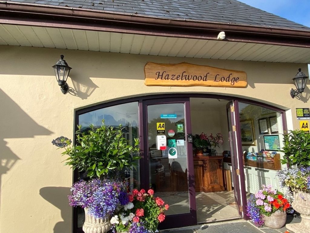 De afbeelding toont de gastvrije entree van Hazelwood Lodge, waar een houten bord trots boven dubbele glazen deuren hangt. Levendige potplanten flankeren de deuropening, vergezeld door twee elegante zwarte lantaarns en een reeks stickers op het glas. | Echt Ierland Vakanties