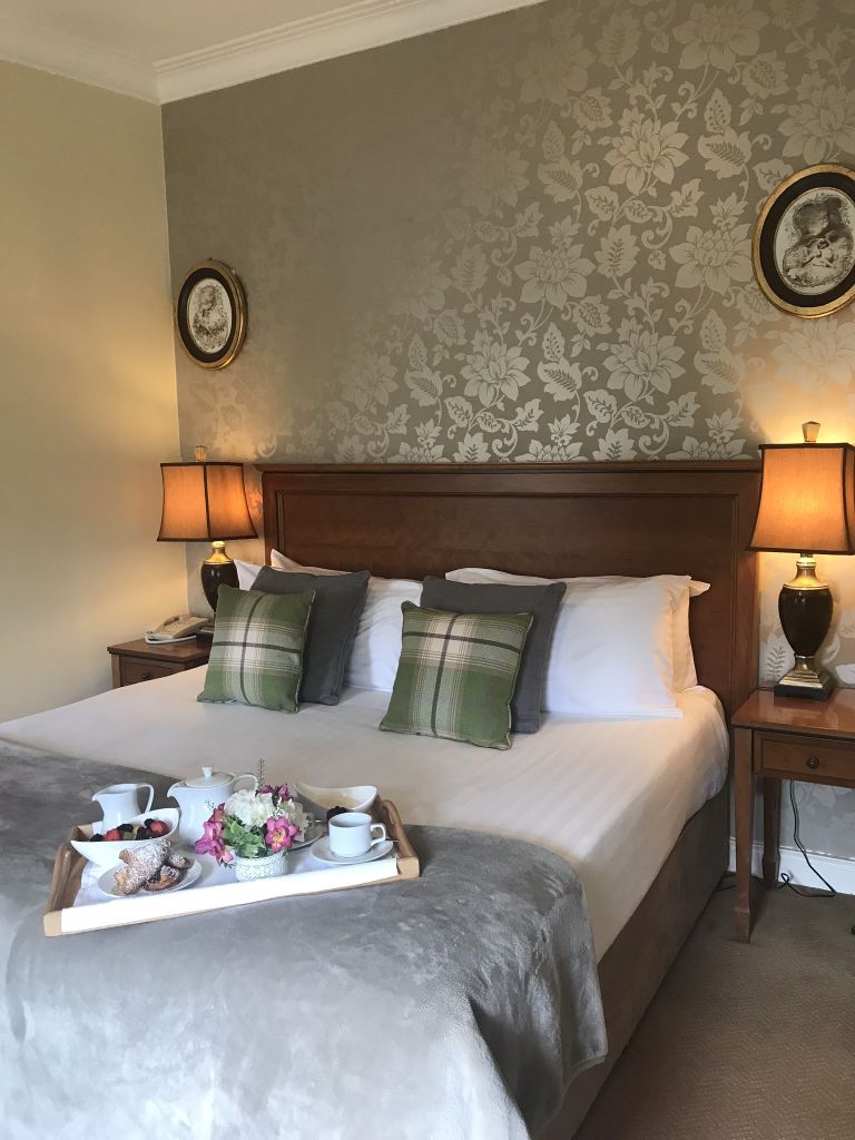 Gezellige slaapkamer met een groot bed met een grijze deken en geruite kussens. Een houten hoofdeinde staat tegen een behang met bloemenmotief. Twee lampen op houten nachtkastjes flankeren het bed. Een ontbijtblad met croissants, drankjes en bloemen staat op het bed. | Echt Ierland Vakanties