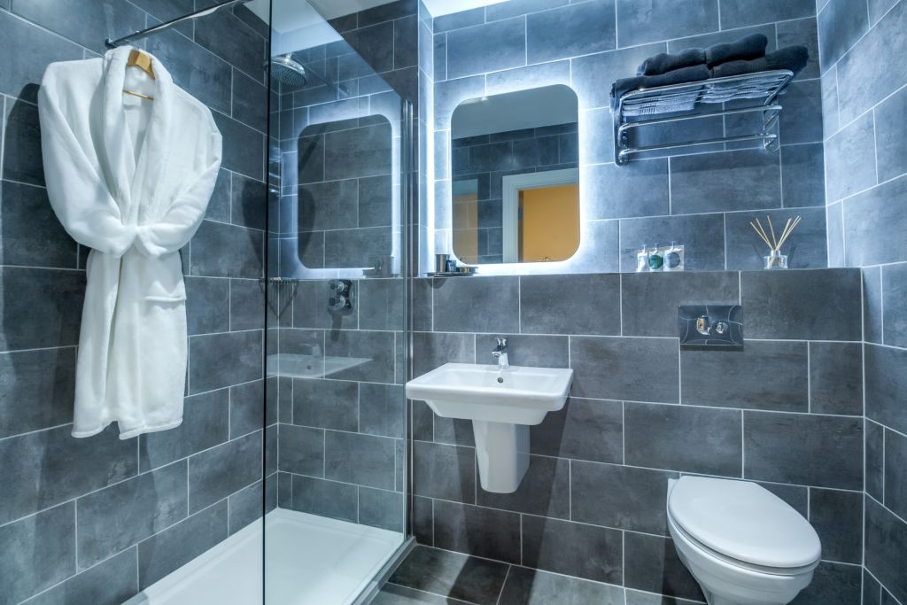 Moderne badkamer met grijze betegelde muren. Kenmerken omvatten een glazen douche met een witte badjas erin, een rechthoekige spiegel boven een minimalistische wastafel, een aan de muur gemonteerd toilet en een metalen rek met zwarte handdoeken. Een kleine geurstokjesverspreider en twee glazen flessen sieren de plank. | Echt Ierland Vakanties