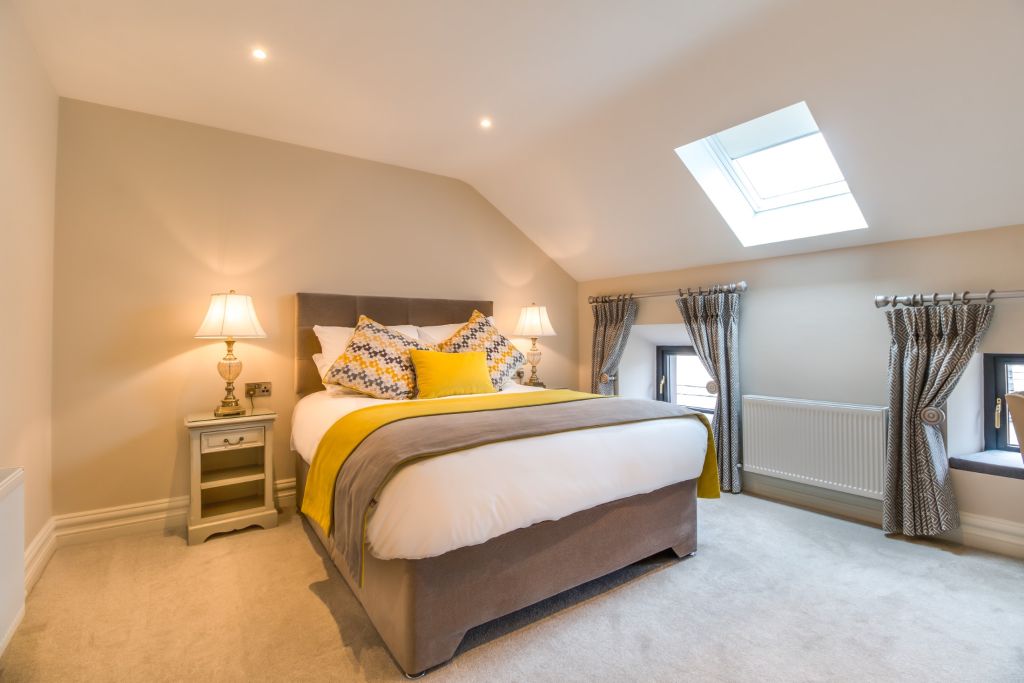 Een gezellige slaapkamer met een beige en schuin plafond. De kamer beschikt over een tweepersoonsbed met een grijs hoofdeinde, wit beddengoed en gele accenten. Twee nachtkastjes hebben elk een lamp met een crèmekleurige kap. Een dakraam en twee kleine ramen met gordijnen laten natuurlijk licht binnen. Vloerbedekking in een neutrale toon. | Echt Ierland Vakanties