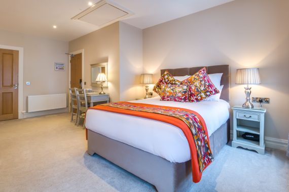 Een netjes ingerichte slaapkamer met een tweepersoonsbed met kleurrijke kussens en een levendig oranje en rood patroon sprei. De kamer wordt zacht verlicht door twee bijpassende tafellampen op grijze nachtkastjes. Een kaptafel met een stoel en spiegel staat tegen de muur. Beige muren en tapijt maken de setting compleet. | Echt Ierland Vakanties