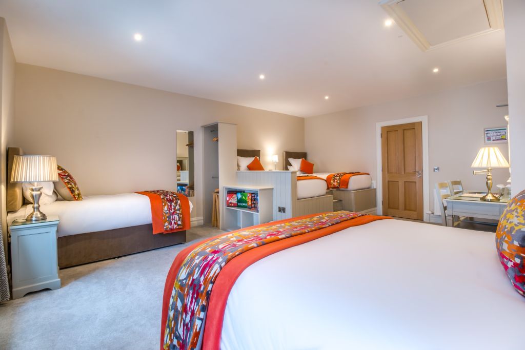 Ruime hotelkamer met drie grote bedden met kleurrijke patchwork-plaids en oranje accenten. Naast elk bed staat een nachtkastje met lampen. Neutrale muren en tapijt creëren een kalmerende sfeer. Een houten deur, stoelen, een kleine tafel en decoratieve wandkunst maken de kamer compleet. | Echt Ierland Vakanties