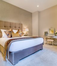 Elegante slaapkamer met een groot bed met een gecapitonneerd hoofdeinde, decoratieve kussens en een kleurrijke sprei. Aan weerszijden van het bed staan stijlvolle lampen op nachtkastjes met spiegels. Een kaptafel met een spiegel en kruk staat tegen de muur en een hoge sierlijke spiegel staat ernaast. Zachte, neutrale tinten domineren. | Echt Ierland Vakanties