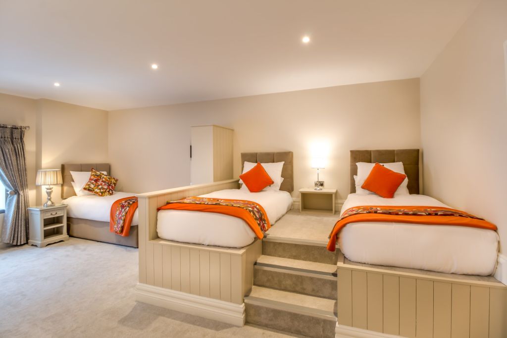 Een slaapkamer met drie eenpersoonsbedden. Twee verhoogde bedden hebben oranje kussens en decoratieve plaids, gescheiden door een centrale lamp op een kleine trede. Het derde bed is lager met een vergelijkbaar ontwerp. Beige muren en tapijt creëren een warme sfeer en een raam met een gordijn is aan de linkerkant. | Echt Ierland Vakanties