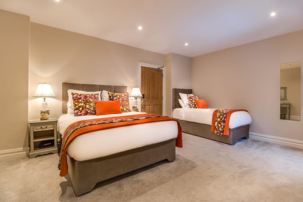 Een gezellige slaapkamer met beige muren heeft een groot bed en een eenpersoonsbed, beide met wit linnen, kleurrijke bloemenspreien en bijpassende kussens. Een houten deur is op de achtergrond, geflankeerd door bedlampjes op nachtkastjes. Een spiegel is op de rechtermuur en zachte verlichting creëert een warme ambiance. | Echt Ierland Vakanties