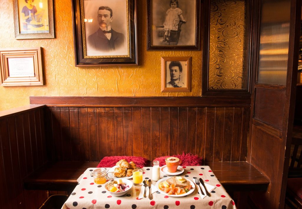 Een gezellige ontbijtsetting in een warm verlichte cabine met houten lambrisering. De tafel is voorzien van een spreiding van gebak, fruit, sap en cappuccino op een wit kleed met rode en zwarte stippen. Vintage portretten hangen aan de getextureerde gouden muren, wat de nostalgische ambiance versterkt. | Echt Ierland Vakanties