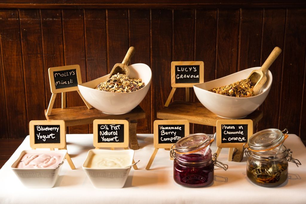 Een ontbijtopstelling met twee grote kommen muesli en granola met houten scheppen op houten standaards. Kleine krijtbordborden labelen ze. Daaronder staan kommen met bosbessenyoghurt en natuurlijke yoghurt, met kleine potjes bessencompote en pruimen in kamille en sinaasappel. | Echt Ierland Vakanties