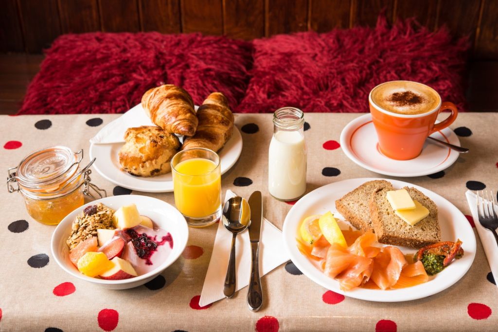 Een ontbijt op een tafelkleed met stippen bestaat uit een pot jam, yoghurt met fruit en granola, croissants, een glas sinaasappelsap, een fles melk, een bord met gerookte zalm, brood en boter en een kop cappuccino. Rode kussens op de achtergrond. | Echt Ierland Vakanties