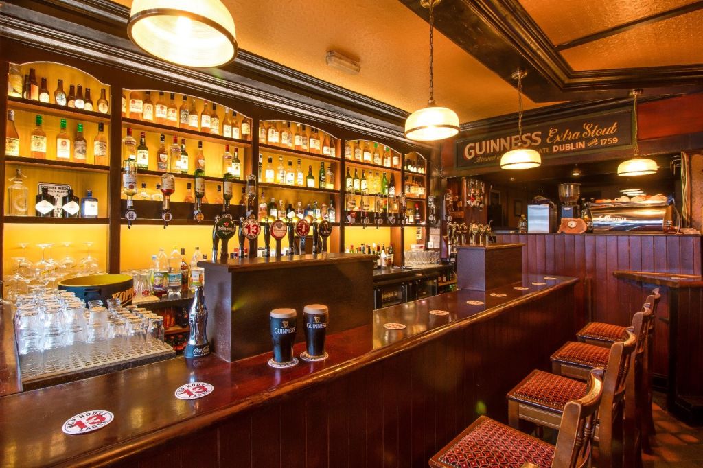 Een gezellig pubinterieur met een goed gevulde bar met verschillende flessen op houten planken. Er zijn biertaps in het midden, waaronder Guinness-opties. Drie barkrukken met rode patroonzittingen staan tegenover de bar. Warme verlichting en een vintage Guinness-bord versterken de uitnodigende sfeer. | Echt Ierland Vakanties