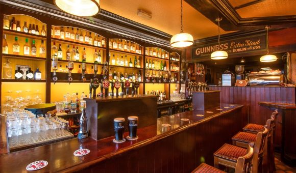 Een gezellig pubinterieur met een goed gevulde bar met verschillende flessen op houten planken. Er zijn biertaps in het midden, waaronder Guinness-opties. Drie barkrukken met rode patroonzittingen staan tegenover de bar. Warme verlichting en een vintage Guinness-bord versterken de uitnodigende sfeer. | Echt Ierland Vakanties