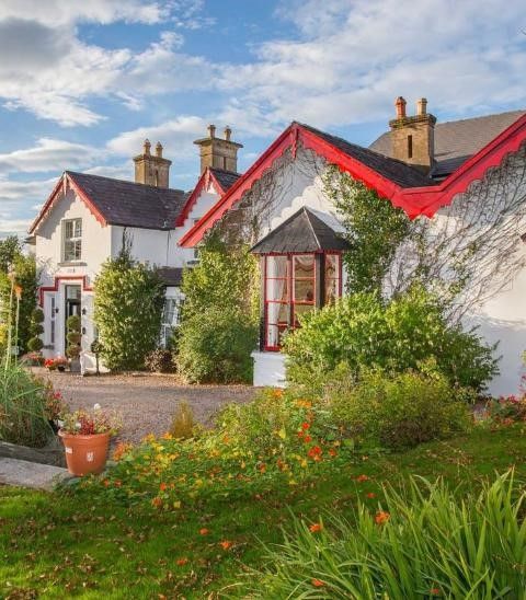 Een charmant wit huisje met levendige rode randen, omgeven door weelderig groen en kleurrijke bloemen. Het gebouw heeft meerdere zadeldaken en een grindpad dat naar de voordeur leidt. Klimop klimt langs de muren en een klassieke lantaarnpaal staat op de grasachtige voorgrond. De lucht is gedeeltelijk bewolkt. | Echt Ierland Vakanties