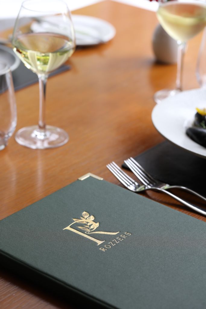 Close-up van een gedekte tafel voor het diner, met een groen menu met een gouden letter "R" en een bladmotief. Twee wijnglazen met witte wijn, een bord met eten, een glas water en bestek zijn zichtbaar op een licht houten oppervlak. De setting is elegant en uitnodigend. | Echt Ierland Vakanties