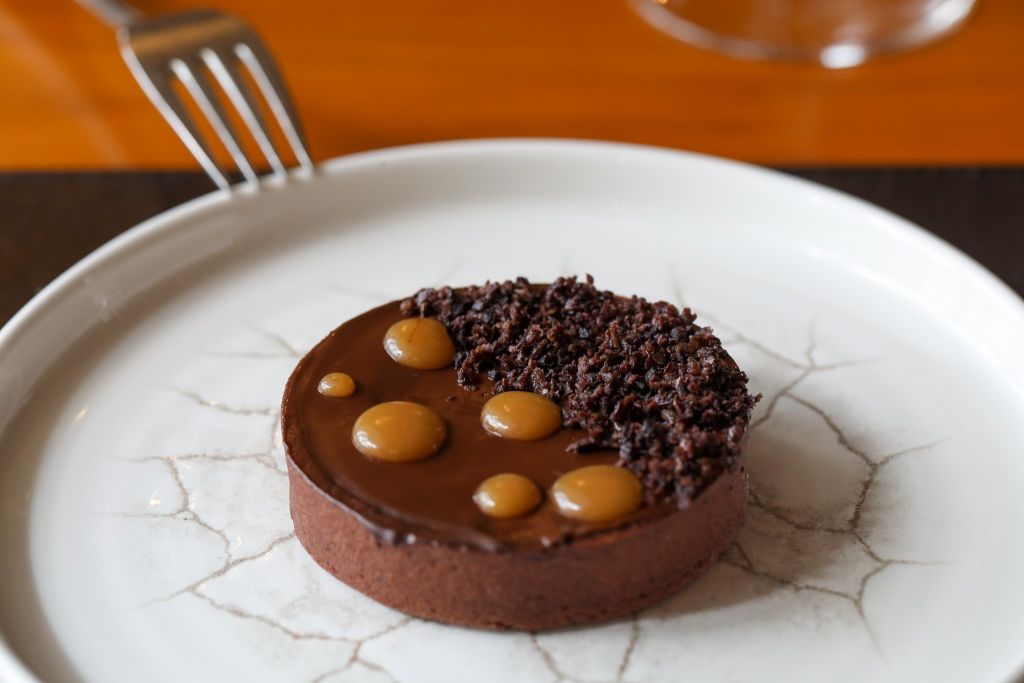 Een ronde chocoladetaart staat op een wit bord met een gebarsten patroon. De taart heeft een gladde chocoladeglazuur met een kruimelig chocolademengsel en glanzende karameldruppels. Linksboven is een vork zichtbaar en op de achtergrond staat een wazig glas op een houten tafel. | Echt Ierland Vakanties