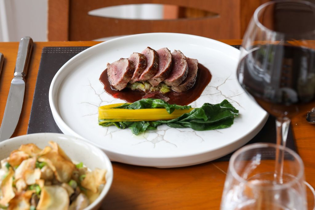 Een gastronomisch gerecht met plakjes, medium-rare rundvlees op een dikke saus op een wit bord. Vergezeld van gebakken groenten en een gele groentestengel. Op de voorgrond een kom chips en een glas rode wijn. Gezet op een houten tafel met bestek en een servet. | Echt Ierland Vakanties