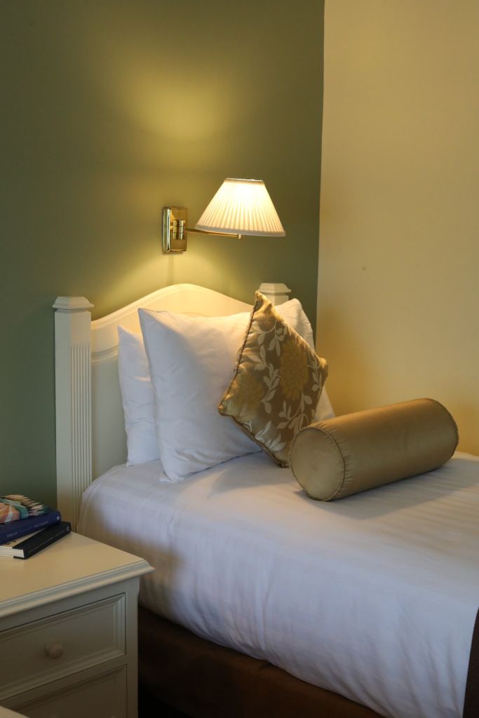 Een knusse slaapkamerhoek met een eenpersoonsbed met een wit hoofdeinde. Het bed is netjes opgemaakt met witte lakens, een decoratief kussen met bloemenmotief en een cilindervormig kussen. Een aan de muur bevestigde lamp met een geplooide kap werpt warm licht op olijf- en crèmekleurige muren. Een nachtkastje bevat tijdschriften. | Echt Ierland Vakanties