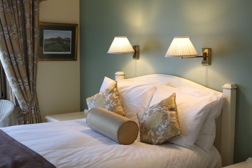 Een gezellige slaapkamer met een wit bed met kussens met bloemenmotief en een cilindervormig kussen. Twee aan de muur gemonteerde lampen met geplooide kappen verlichten de ruimte. Een ingelijst landschapsschilderij en gordijnen met patronen sieren de lichtgroene muren, wat een serene sfeer creëert. | Echt Ierland Vakanties