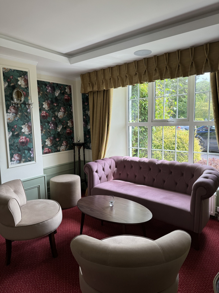 Een gezellige zithoek met een roze gecapitonneerde bank, twee beige gestoffeerde stoelen en een ronde houten tafel. Er staat een kleine kaars op de tafel. De kamer heeft bloemenbehang, gouden gordijnen die een groot raam met uitzicht op de tuin omlijsten en een rode vloerbedekking. | Echt Ierland Vakanties