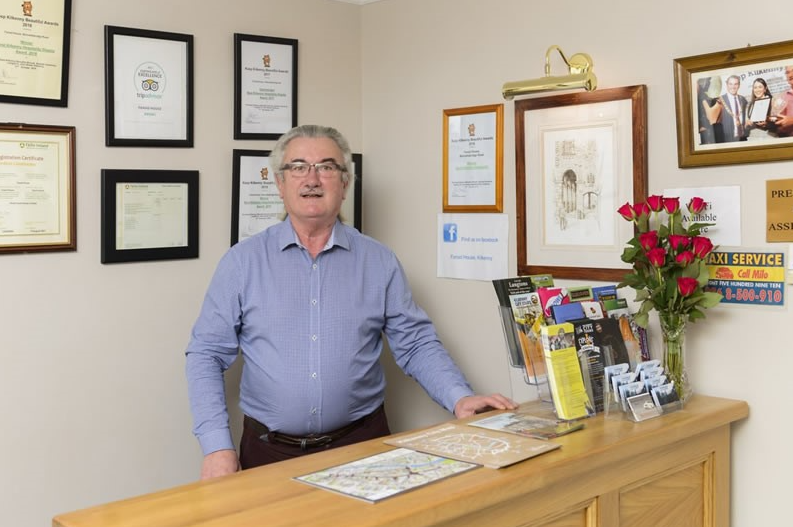 Een grijsharige man met bril staat achter een houten receptie met brochures, kaarten en rode rozen in een vaas. Aan de muur achter hem hangen ingelijste certificaten, prijzen en foto's. De omgeving lijkt een gastvrij kantoor of klein bedrijf te zijn. | Echt Ierland Vakanties