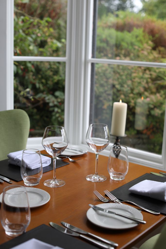 Een houten tafel is elegant gedekt met lege wijnglazen, waterglazen en witte borden. Zilverwerk is netjes gerangschikt op zwarte placemats, vergezeld door gevouwen witte servetten. Een brandende kaars staat bij het raam, met weelderig groen zichtbaar buiten. | Echt Ierland Vakanties