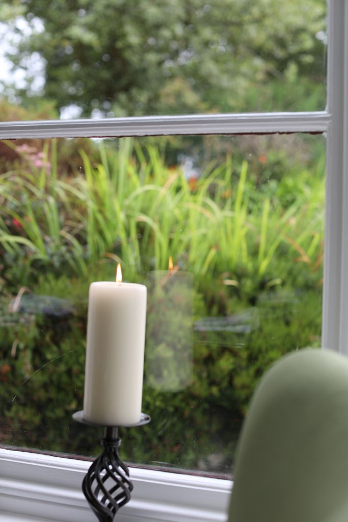 Een brandende witte pilaarkaars op een zwarte houder staat op een vensterbank. Het raam kijkt uit op een weelderige tuin met groene planten en struiken. Het tafereel is kalm en sereen, met wazig gebladerte op de achtergrond zichtbaar door het heldere glas. | Echt Ierland Vakanties