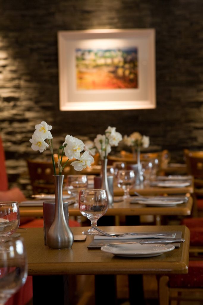 Het restaurantinterieur van Hotel Granville beschikt over netjes opgestelde tafels, versierd met elegante witte bloemen in zilveren vazen. Elke tafel is gedekt met wijnglazen, witte servetten en borden, terwijl een zacht verlicht schilderij op de stenen muur een gezellige ambiance toevoegt aan deze verfijnde hotel-eetervaring. | Echt Ierland Vakanties