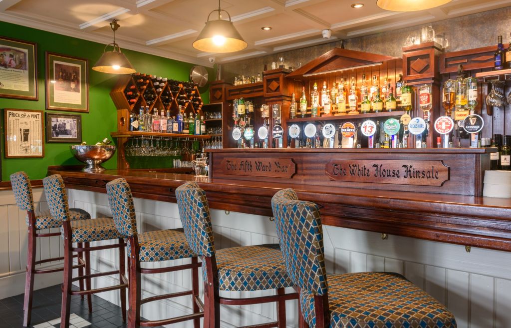 Een gezellig pubinterieur dat doet denken aan het Hotel Granville, met een lange houten bar met zes barkrukken met patronen. Versierd met talloze biertaps en flessen op planken, gloeit er warme verlichting van hanglampen boven. De groene muren tonen ingelijste foto's en een wijnrek op de achtergrond. | Echt Ierland Vakanties