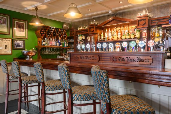Een gezellig pubinterieur dat doet denken aan het Hotel Granville, met een lange houten bar met zes barkrukken met patronen. Versierd met talloze biertaps en flessen op planken, gloeit er warme verlichting van hanglampen boven. De groene muren tonen ingelijste foto's en een wijnrek op de achtergrond. | Echt Ierland Vakanties