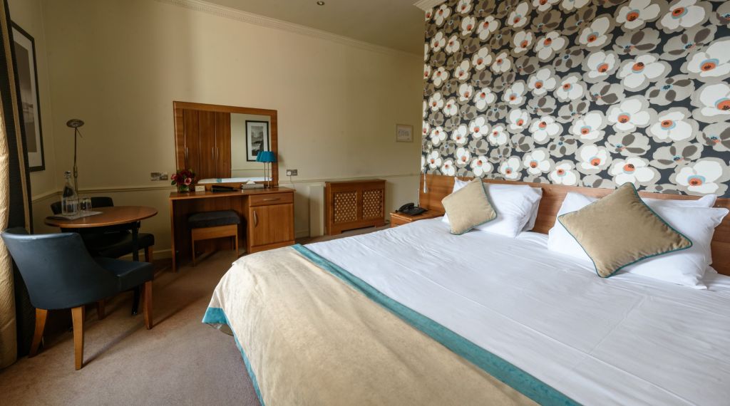 De Hotel Granville kamer heeft een kingsize bed met wit linnen en zachte kussens. Achter het bed siert een groot bloemenpatroon de muur. Een kleine ronde tafel, twee stoelen en een ladekast met een spiegel sieren de vloerbedekking, allemaal omgeven door zacht licht voor een uitnodigende ambiance. | Echt Ierland Vakanties