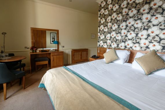 De Hotel Granville kamer heeft een kingsize bed met wit linnen en zachte kussens. Achter het bed siert een groot bloemenpatroon de muur. Een kleine ronde tafel, twee stoelen en een ladekast met een spiegel sieren de vloerbedekking, allemaal omgeven door zacht licht voor een uitnodigende ambiance. | Echt Ierland Vakanties