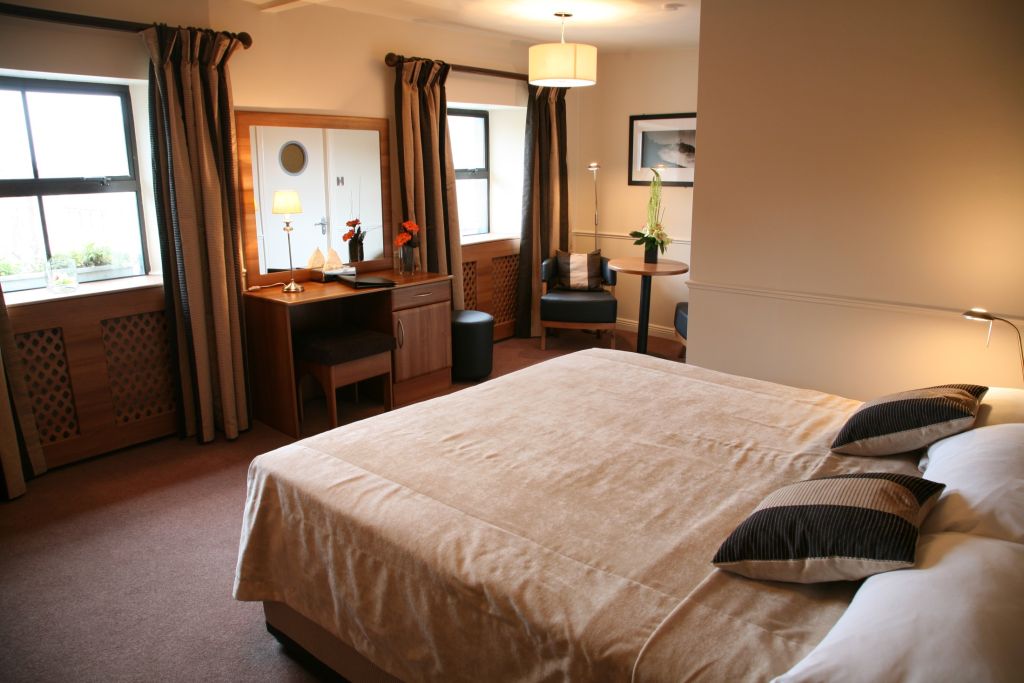 Een gezellige hotelkamer met een groot beige bed, twee bijpassende kussens en bruine accenten. Een houten kaptafel met een spiegel en lamp staat tegen de muur. Twee ramen met bruine gordijnen verwelkomen natuurlijk licht. Een kleine tafel met een plant en twee stoelen sieren de hoek in echte Granville-stijl. | Echt Ierland Vakanties