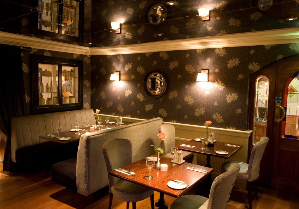 Een gezellig restaurantinterieur in Hotel Granville, met gedimde verlichting. Geniet van grijze, met kussens beklede zitplaatsen en houten tafels versierd met wijnglazen en servies. Behang met bloemenmotief en kleine ronde spiegels sieren de muren, terwijl wandlampen zachte verlichting bieden. | Echt Ierland Vakanties