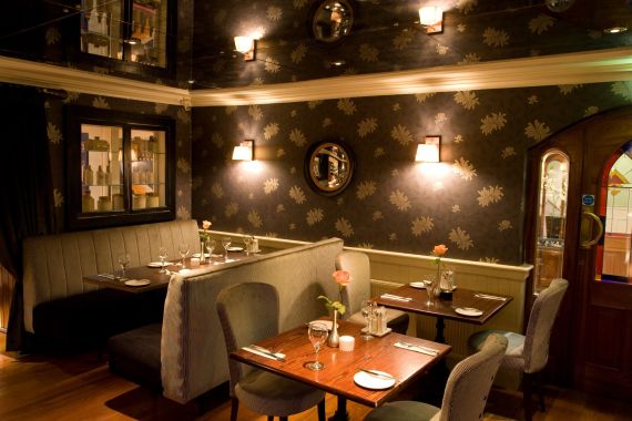 Een gezellig restaurantinterieur in Hotel Granville, met gedimde verlichting. Geniet van grijze, met kussens beklede zitplaatsen en houten tafels versierd met wijnglazen en servies. Behang met bloemenmotief en kleine ronde spiegels sieren de muren, terwijl wandlampen zachte verlichting bieden. | Echt Ierland Vakanties