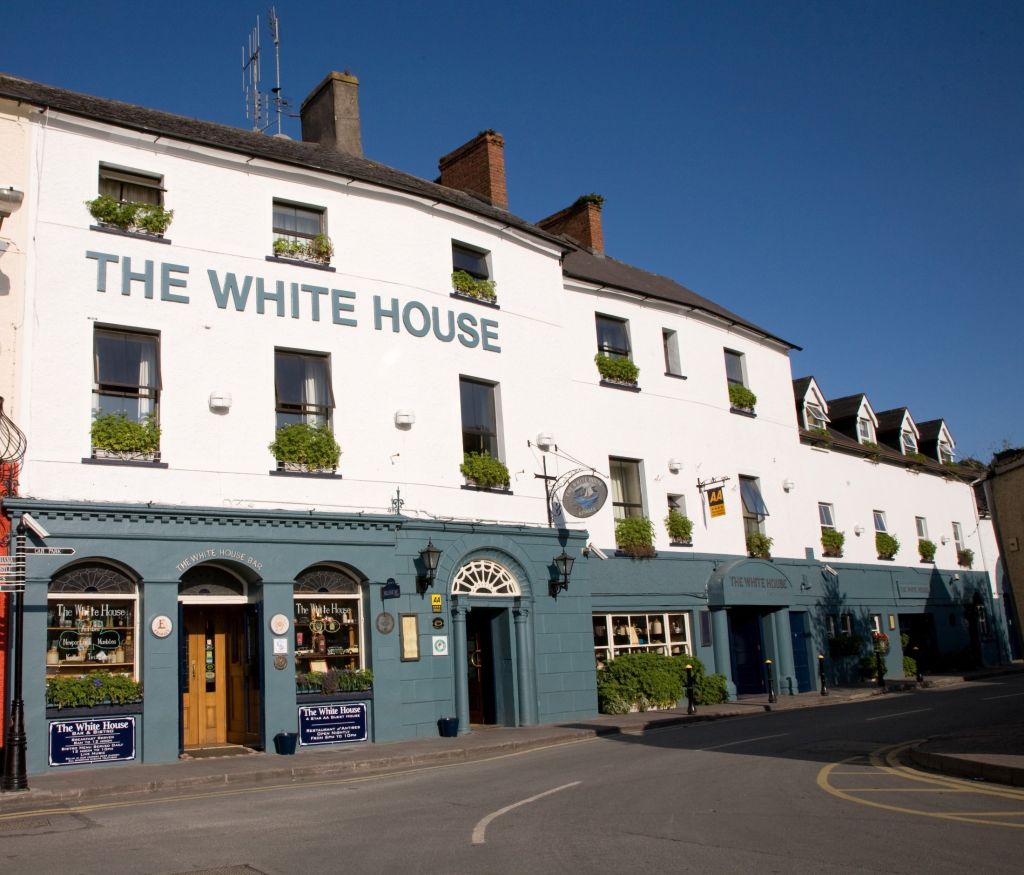 Hoekaanzicht van een gebouw van drie verdiepingen met "The White House Hotel" erop geschilderd. Het onderste gedeelte is blauwgroen en herbergt een pub met ronde borden en krijtborden. De bovenste verdiepingen zijn wit met kleine ramen en bloembakken. Het staat tegen een helderblauwe lucht, met een bochtige weg op de voorgrond. | Echt Ierland Vakanties