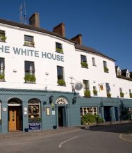Hoekaanzicht van een gebouw van drie verdiepingen met "The White House Hotel" erop geschilderd. Het onderste gedeelte is blauwgroen en herbergt een pub met ronde borden en krijtborden. De bovenste verdiepingen zijn wit met kleine ramen en bloembakken. Het staat tegen een helderblauwe lucht, met een bochtige weg op de voorgrond. | Echt Ierland Vakanties