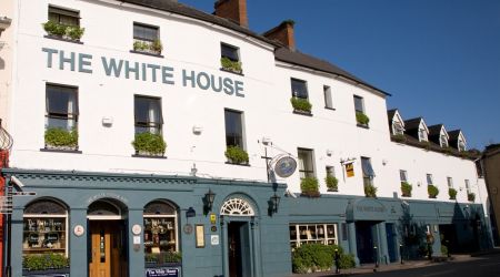Hoekaanzicht van een gebouw van drie verdiepingen met "The White House Hotel" erop geschilderd. Het onderste gedeelte is blauwgroen en herbergt een pub met ronde borden en krijtborden. De bovenste verdiepingen zijn wit met kleine ramen en bloembakken. Het staat tegen een helderblauwe lucht, met een bochtige weg op de voorgrond. | Echt Ierland Vakanties
