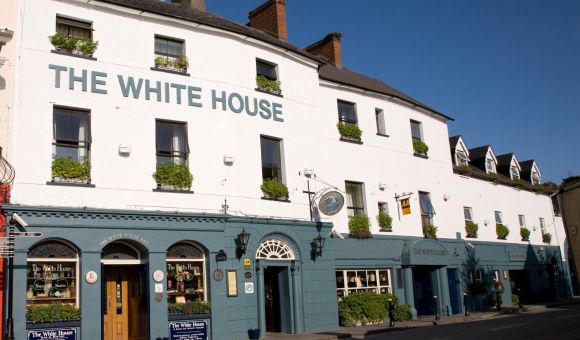 Hoekaanzicht van een gebouw van drie verdiepingen met "The White House Hotel" erop geschilderd. Het onderste gedeelte is blauwgroen en herbergt een pub met ronde borden en krijtborden. De bovenste verdiepingen zijn wit met kleine ramen en bloembakken. Het staat tegen een helderblauwe lucht, met een bochtige weg op de voorgrond. | Echt Ierland Vakanties